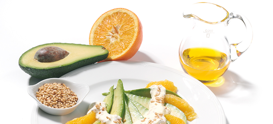 Eine halbe Avocado und eine halbe Orange liegen neben einem Teller mit geschnittenen Avocados und Orangen, neben einer Kanne mit Öl aus guten Fetten, auf weißem Hintergrund.